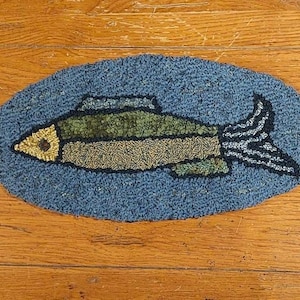 Può includere: Un tappeto a forma di pesce blu e verde con un occhio giallo, su una superficie di legno.