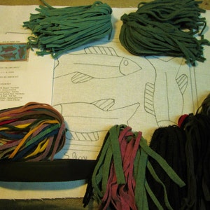 May include: A needlepoint kit featuring a design of three fish with a black outline on white canvas. The kit includes a variety of colored felt strips for embroidery, a black felt strip, and instructions.