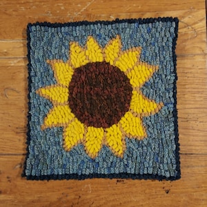 Op de afbeelding: Een blauw en geel kleed met een zonnebloemdessin. De zonnebloem heeft een bruin hart en gele bloemblaadjes.