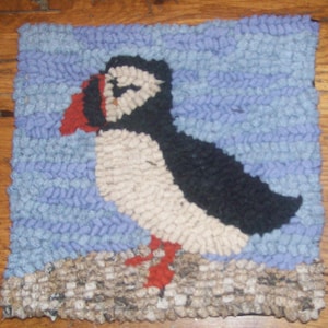 Puede incluir: Una alfombra hecha a mano que representa un frailecillo de pie sobre una roca. El frailecillo es blanco con la espalda negra y el pico naranja. El fondo es de color azul claro.