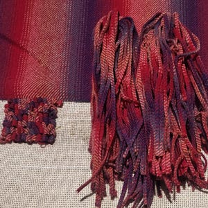 May include: A woven fabric with a fringe of red, purple, and pink yarn. The fabric has a striped pattern and a small woven square in the corner.