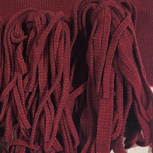 May include: Close-up of a burgundy textile with a fringe detail. The fringe is made of long, thick strands of the same burgundy material, creating a textured and layered effect. The fabric appears soft and possibly knitted or woven.