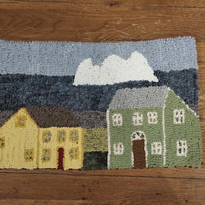 Op de afbeelding: Handgetuft tapijt met een schilderachtige dorpsscène. Het tapijt toont twee huizen, een gele en een groene, tegen een achtergrond van een bewolkte lucht. De huizen hebben gedetailleerde ramen en deuren, en het tapijt ligt op een houten vloer.