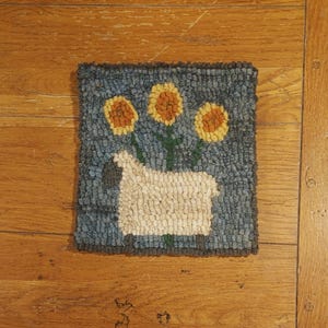 May include: A square, hand-hooked rug depicting a white sheep with a grey head and three yellow sunflowers against a blue backdrop. The rug has a dark blue border and is set on a wooden surface. The rug is approximately 30 cm square.
