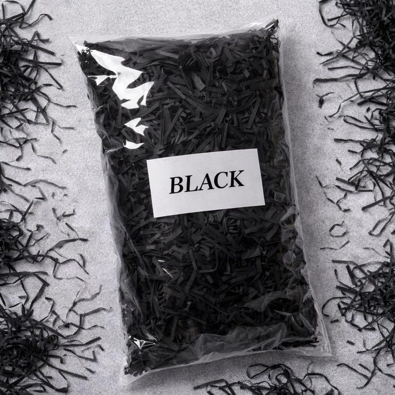 May include: A clear plastic bag filled with black shredded paper, with the word "BLACK" on a white label. The shredded paper is scattered around the bag on a gray surface.