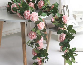 Könnte beinhalten: Eine Girlande aus rosa Rosen und grünen Blättern drapiert über einem weißen Tisch. Die künstlichen Blumen sind in voller Blüte, mit mehreren Rosen und Blättern. Die Girlande ist ein dekorativer Artikel, geeignet für Hochzeiten, Partys oder Heimdekoration.