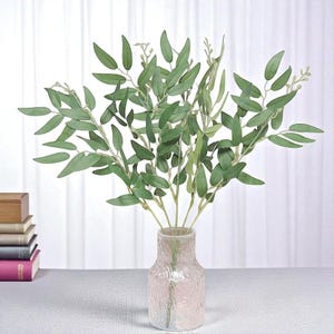 May include: A bunch of artificial eucalyptus leaves in a textured glass vase. The leaves are a vibrant green, with long, slender shapes. The vase is a light pink colour, and the background is a white curtain.