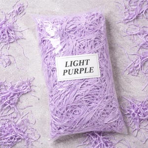 May include: A clear plastic bag filled with light purple shredded paper. The bag has a white label that reads "LIGHT PURPLE". Some shredded paper is scattered around the bag on a gray surface. This is a decorative packaging material.