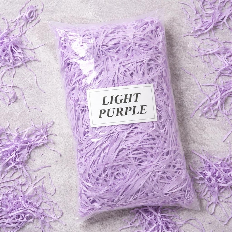 May include: A clear plastic bag filled with light purple shredded paper. The bag has a white label that reads "LIGHT PURPLE". Some shredded paper is scattered around the bag on a gray surface. This is a decorative packaging material.