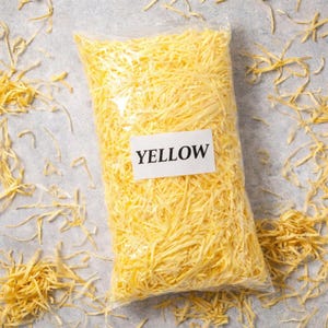 May include: A clear plastic bag filled with yellow shredded paper, with the word "YELLOW" on a white label. The paper is scattered around the bag on a gray surface.