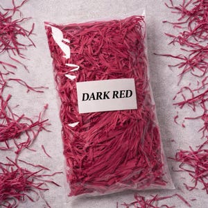 May include: A clear plastic bag filled with dark red shredded paper. A white label with the words "DARK RED" is attached to the bag. The shredded paper is scattered around the bag on a gray surface. This is a decorative packaging material.