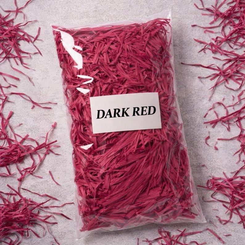 May include: A clear plastic bag filled with dark red shredded paper. A white label with the words "DARK RED" is attached to the bag. The shredded paper is scattered around the bag on a gray surface. This is a decorative packaging material.