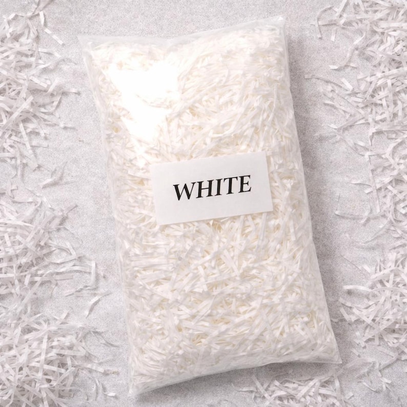 May include: A clear plastic bag filled with white shredded paper. A white rectangular label with the word "WHITE" is affixed to the bag. The shredded paper is scattered around the bag on a light gray surface, creating a clean, minimalist aesthetic.