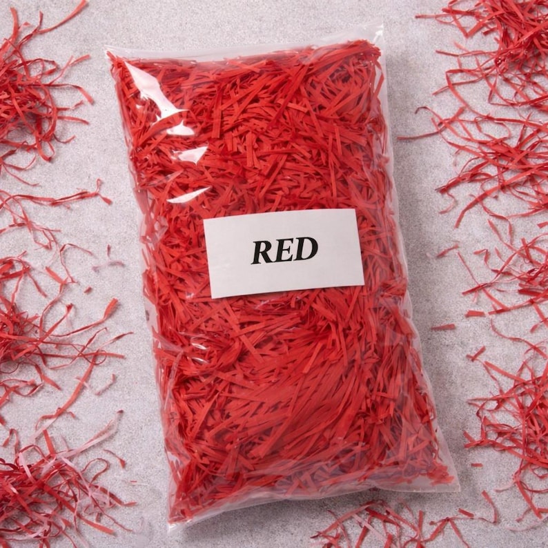 May include: A clear plastic bag filled with bright red shredded paper. A white label with the word "RED" is affixed to the front of the bag. The shredded paper is scattered around the bag on a light gray surface.