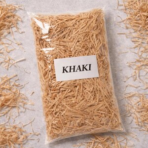 May include: A clear plastic bag filled with khaki-colored shredded paper. A white label with the word "KHAKI" in black capital letters is affixed to the front of the bag. Loose paper shreds are scattered around the bag on a gray surface.