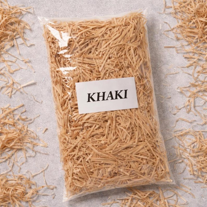 May include: A clear plastic bag filled with khaki-colored shredded paper. A white label with the word "KHAKI" in black capital letters is affixed to the front of the bag. Loose paper shreds are scattered around the bag on a gray surface.