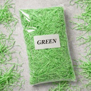 May include: A clear plastic bag filled with bright green shredded paper. A white label on the bag reads "GREEN" in bold, black capital letters. Loose paper is scattered around the bag on a gray surface.