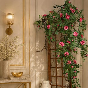 Könnte beinhalten: Ein hohes Holzgitter, das mit künstlichen Kletterrosenranken mit leuchtend rosa Blüten und grünen Blättern bedeckt ist. Daneben befinden sich eine goldene Wandleuchte, eine Marmorvase mit weißen Zweigen und eine goldene Schale auf einem Tisch.