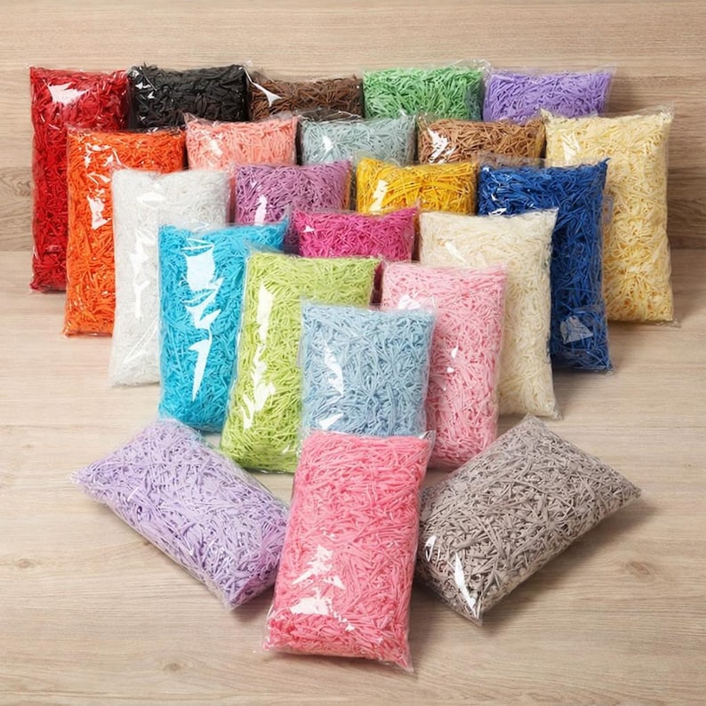 May include: A collection of colorful shredded paper in clear plastic bags. The assortment includes shades of red, orange, black, brown, green, purple, pink, yellow, blue, and white. Ideal for gift packaging or craft projects.