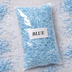May include: A clear plastic bag filled with light blue shredded paper. A white label with the word "BLUE" is attached to the bag. Some shredded paper is scattered around the bag on a light gray surface.