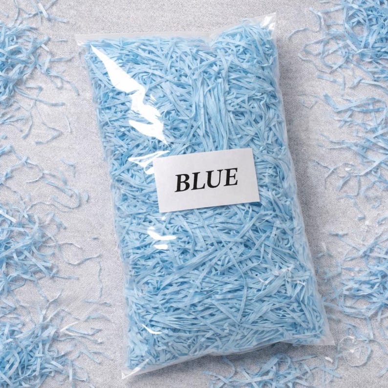 May include: A clear plastic bag filled with light blue shredded paper. A white label with the word "BLUE" is attached to the bag. Some shredded paper is scattered around the bag on a light gray surface.