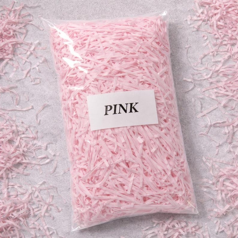 May include: A clear plastic bag filled with shredded pink paper. A white label on the bag reads "PINK" in black capital letters. Loose pink paper shreds are scattered around the bag on a light gray surface.