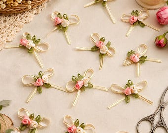 May include: A collection of small decorative bows made from cream-colored ribbon with gold metallic edges. Each bow is embellished with two miniature fabric roses in shades of pink and cream, accented by small green fabric leaves.