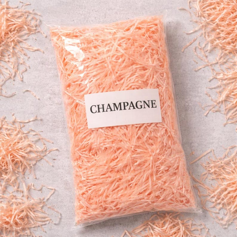 May include: A clear plastic bag filled with peach-colored paper shreds. A white label with the word "CHAMPAGNE" is affixed to the bag. The shreds are scattered around the bag on a light gray surface.