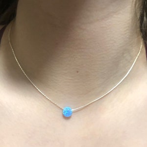May include: A delicate necklace featuring a small, round, light blue opal pendant. The pendant is suspended from a thin, silver-colored chain. The necklace is shown against a skin-toned background.