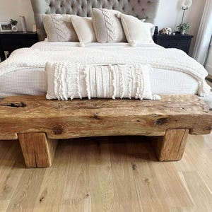 May include: A rustic wooden bench with a natural, weathered appearance. The bench is made from a thick, solid piece of wood, supported by two sturdy wooden legs. The bench is placed at the foot of a bed with white bedding and decorative pillows.