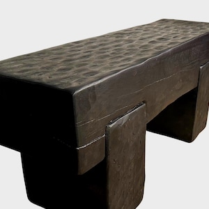May include: A dark brown wooden bench with a textured top and a simple, sturdy design. The bench has two legs on each side, creating a rectangular shape.