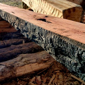 May include: A close-up of a large, weathered wooden beam with a dark, charred surface. The wood has a reddish-brown color and shows signs of age and use.