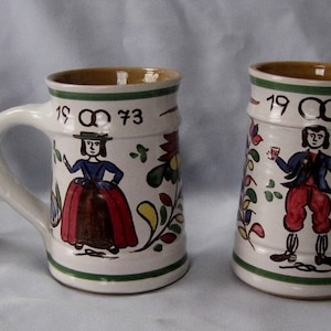May include: Two white ceramic mugs with green, yellow, and red painted designs. The mugs have the date "19 73" and a ring symbol painted on them. The mugs are decorated with painted figures of a man and a woman.