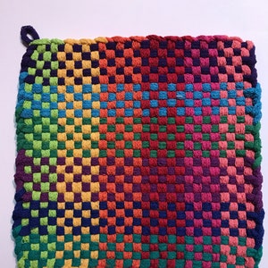 May include: A colorful woven pot holder with a rainbow pattern. The pot holder is made of thick yarn and has a checkered design.