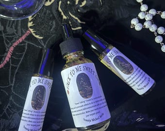 May include: Three glass bottles of essential oils with the text "Talk to Me Sweet Oil" and "Sweet Talking" on the labels. The bottles are on a dark, patterned surface, with a rosary and wine glasses in the background. The oils are from Vemaja Wicked Herbs.