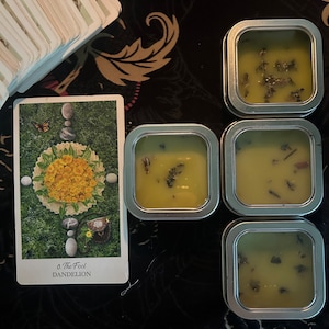 May include: Four small, round, silver tins filled with a green, herbal salve. The tins are arranged in a square shape on a black background. A tarot card with a green background and a yellow dandelion flower is visible in the upper left corner of the image. The card reads "0. The Fool, Dandelion."