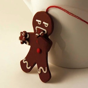 May include: A brown gingerbread man tea bag holder with white icing details and a red button, attached to a white mug with a red string. The gingerbread man has an angry expression. The mug is white.
