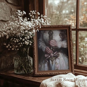 May include: A framed portrait of a person holding a sword with a pink ribbon. The portrait is in a brown frame and is sitting on a wooden surface. The text "SLEEPY ROOK HOLLOW" is visible below the frame.