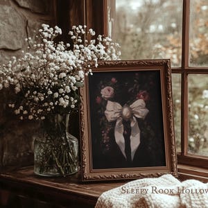 May include: A framed artwork featuring a white bow with a cameo brooch, set against a dark floral background. The artwork is displayed on a wooden surface near a window, alongside a vase of white flowers. The text "Sleepy Rook Hollow" is visible at the bottom.