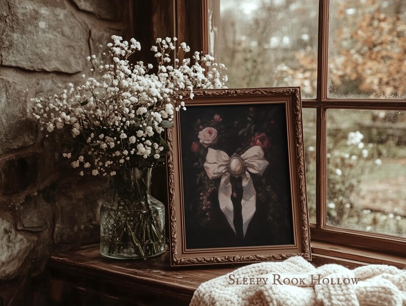 May include: A framed artwork featuring a white bow with a cameo brooch, set against a dark floral background. The artwork is displayed on a wooden surface near a window, alongside a vase of white flowers. The text "Sleepy Rook Hollow" is visible at the bottom.