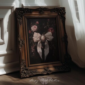 May include: A framed painting with a dark background, featuring a white bow with a central brooch and roses. The ornate, gold-toned frame adds a vintage aesthetic. The painting is leaning against a white wall.