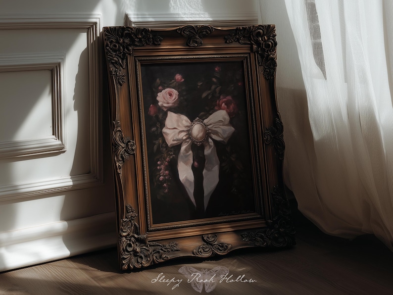 May include: A framed painting with a dark background, featuring a white bow with a central brooch and roses. The ornate, gold-toned frame adds a vintage aesthetic. The painting is leaning against a white wall.
