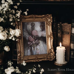 May include: A framed portrait of a woman in a white dress holding a sword. The frame is gold and ornate. The portrait is set on a shelf with books and a candle.