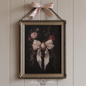 May include: A framed artwork featuring a large white bow with a cameo in the center, surrounded by roses and foliage. The artwork is in an ornate gold frame and is suspended by a chain. A matching pink bow is tied above the frame.