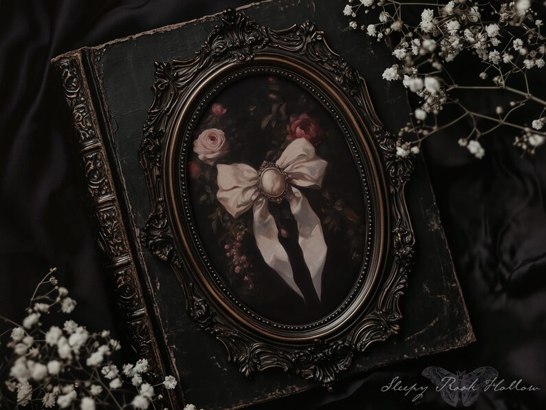 May include: An antique-style framed oval print featuring a bow with a pearl-like center, roses, and a dark background. The frame is ornate and dark, resting on a dark green, aged book. White flowers are in the foreground.