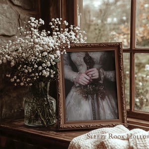 K&ouml;nnte beinhalten: Ein gerahmtes Portr&auml;t einer Frau in einem wei&szlig;en Kleid, die einen Blumenstrau&szlig; h&auml;lt. Das Portr&auml;t ist in einem goldenen Rahmen und steht auf einer Holzoberfl&auml;che. Der Text "SLEEPY ROOK HOLLOW" ist unterhalb des Rahmens sichtbar.