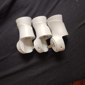 May include: Three white air freshener plug-in units. Each unit has a curved design with a textured pattern on the side. The plug-in prongs are visible on one of the units. The units are arranged on a dark surface.