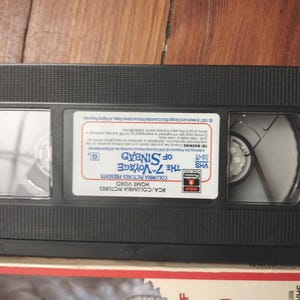 Peut inclure: Une cassette VHS noire avec l'inscription "The 7th Voyage of Sinbad" de Columbia Pictures. L'étiquette blanche porte du texte rouge et bleu. La cassette est posée sur une surface en bois.