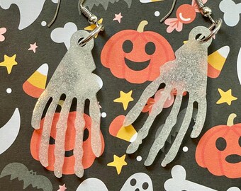 May include: A pair of silver skeleton hand earrings with a glittery finish. The earrings are on a black background with a Halloween theme.