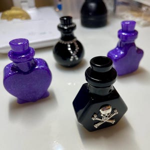 May include: An assortment of decorative potion bottles in various shapes and colors. The bottles are black and purple, with glitter accents. One bottle features a skull and crossbones design, while another has a heart shape. The bottles are likely for decorative purposes.